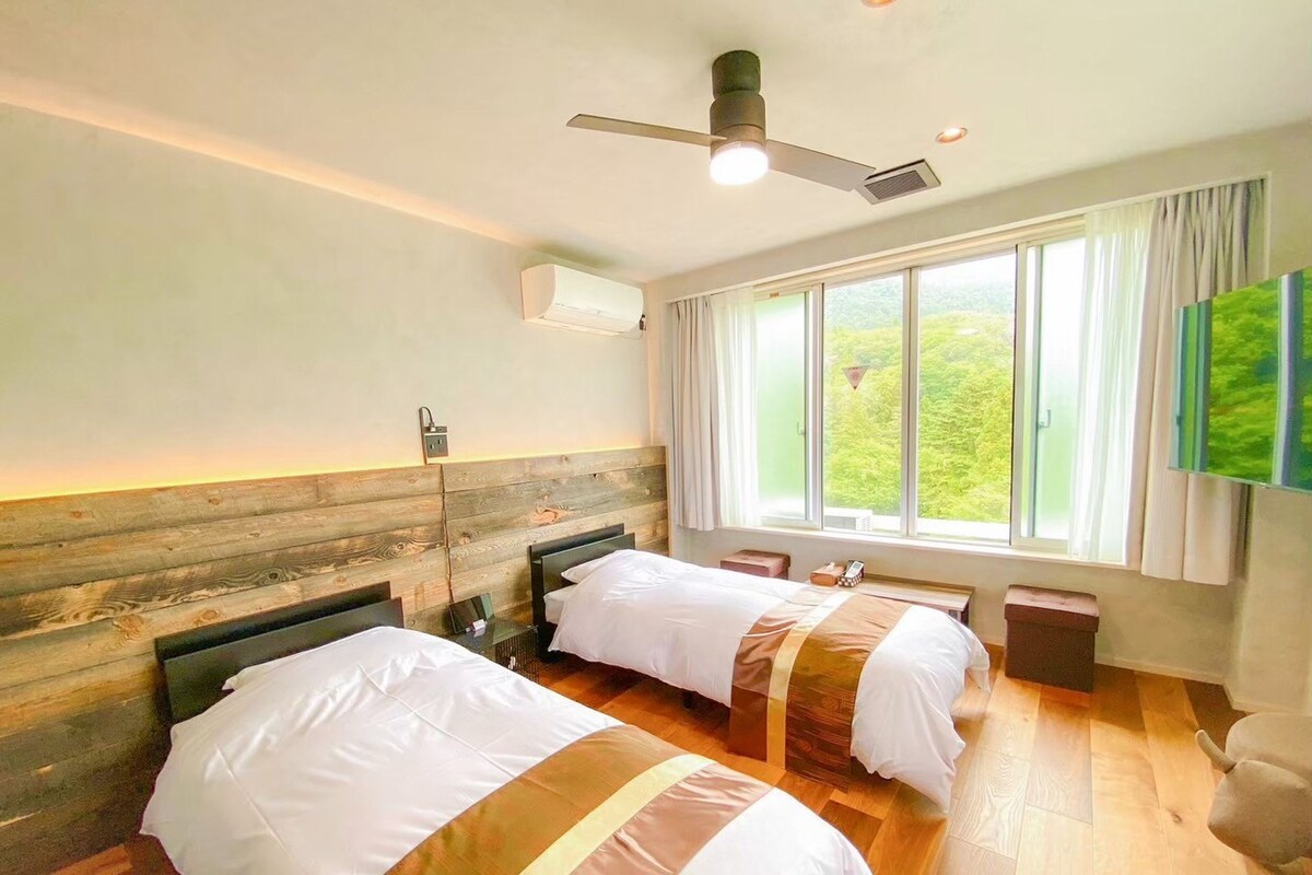 A hotel room with two beds, a ceiling fan, an air conditioning unit, a window with a view, and a flat-screen TV mounted on the wall.