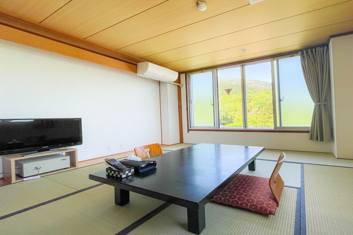 A room with a large window, a flat-screen TV, a table, and traditional tatami flooring.