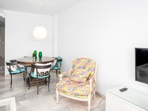 A modern living room with a round dining table, two chairs, a floral armchair, a flat-screen TV, and a small cabinet. - Los Arcos by Interhome (Torremolinos)