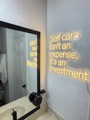 A bathroom with a mirror, a sink, and a wall-mounted light fixture displaying motivational text. - Modern 4BR, Patio, BBQ Grill and Theater— Perfect For (Grand Prairie)