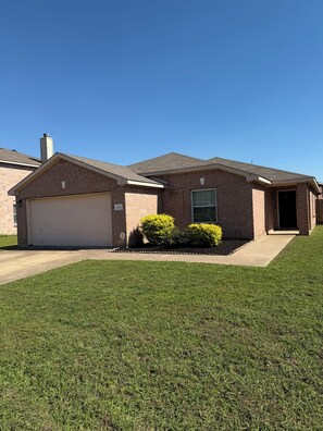A two-story brick house with a garage, a front door, and a small front yard with grass and shrubs. - Modern 4BR, Patio, BBQ Grill and Theater— Perfect For (Grand Prairie)
