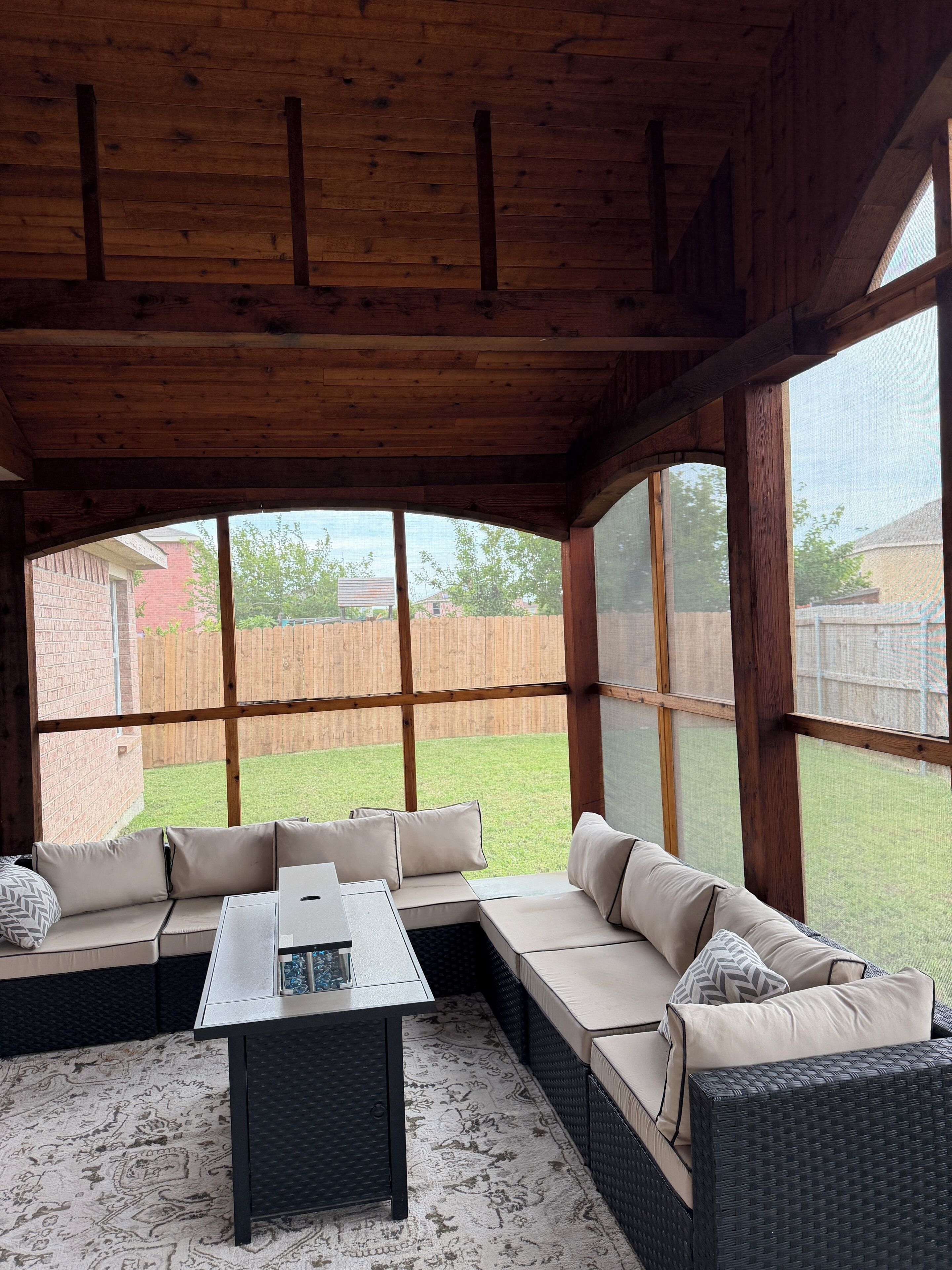 A covered patio area with a sectional sofa, a small table, and large windows offering a view of a backyard.