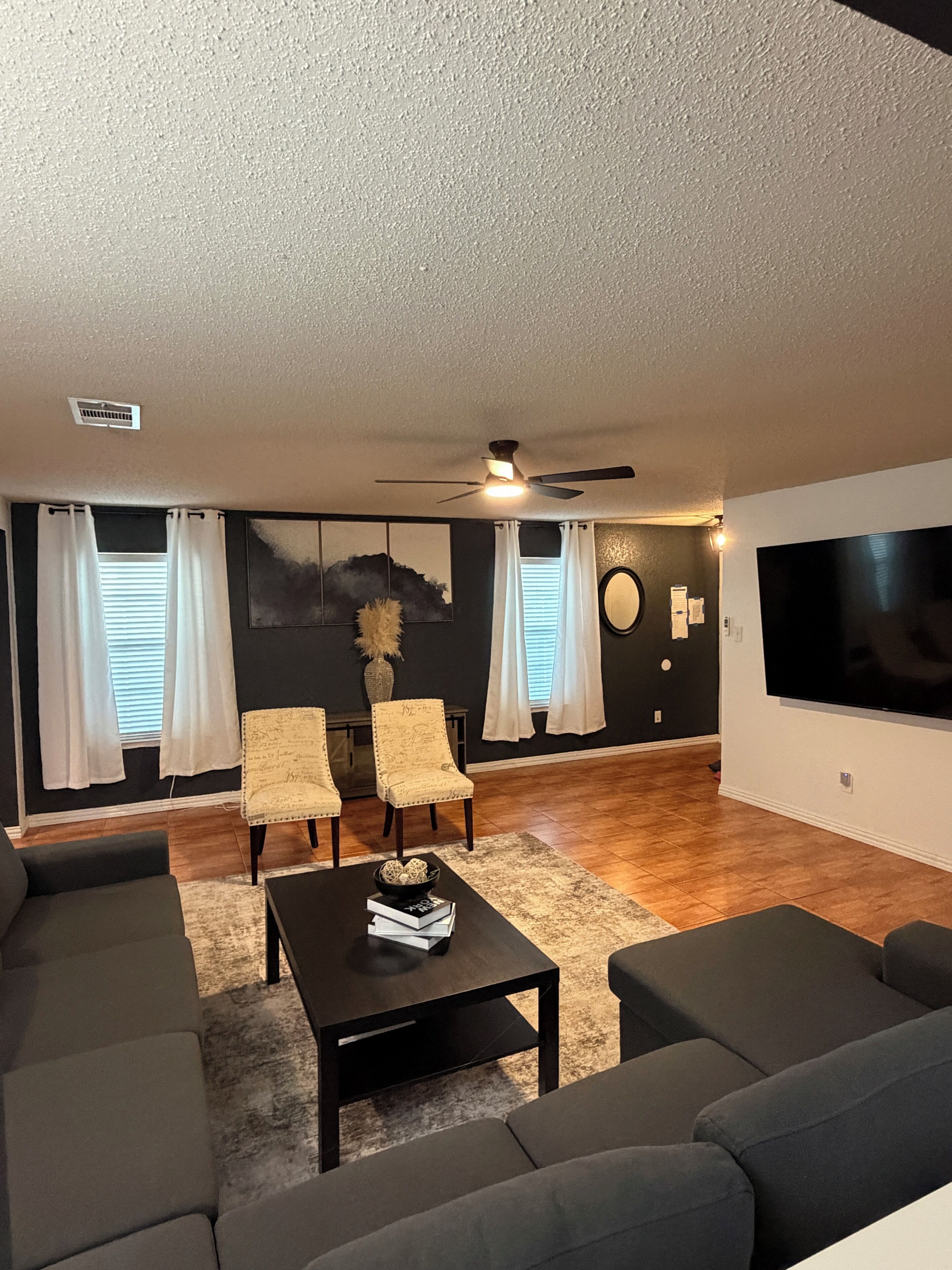 A living room with a sofa set, a coffee table, two armchairs, a ceiling fan, and a flat-screen TV.