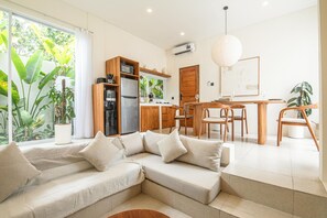 A modern living room with a sectional sofa, a dining area with wooden chairs, and a kitchen with a refrigerator and microwave. - Brand New 2br Uluwatu Villa 2 Mins to Beach (Pecatu)