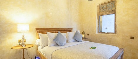 Une chambre à coucher avec un lit en bois, du linge de lit blanc, une lampe de chevet et une fenêtre ornée d’un cadre décoratif.