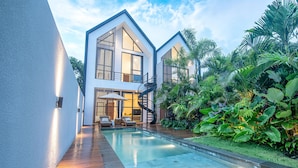 Une maison moderne à deux étages avec une piscine, entourée d’une végétation tropicale luxuriante.