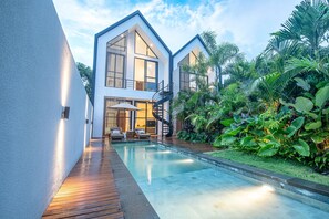 Ein modernes, zweistöckiges Haus mit Swimmingpool, umgeben von üppiger tropischer Vegetation.