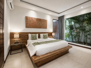Ein modernes Schlafzimmer mit einem großen Bett, einem Holz-Kopfteil, einem Wandbild, einem Nachttisch mit Lampe und Blick auf Zimmerpflanzen durch ein Fenster.