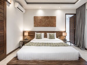A modern bedroom with a large bed, wooden headboard, two bedside tables with lamps, a wall-mounted air conditioner, and a large abstract painting. - Splendid Villa 3Br Center Seminyak (Keroboken Kelod)