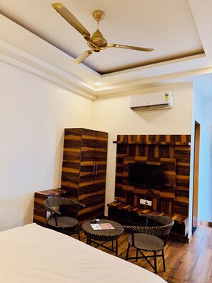 A hotel room with a bed, a desk, two chairs, a television mounted on a wooden wall, a ceiling fan, and an air conditioning unit. - THE CARNATION HOTEL & RESORT (Jaipur)
