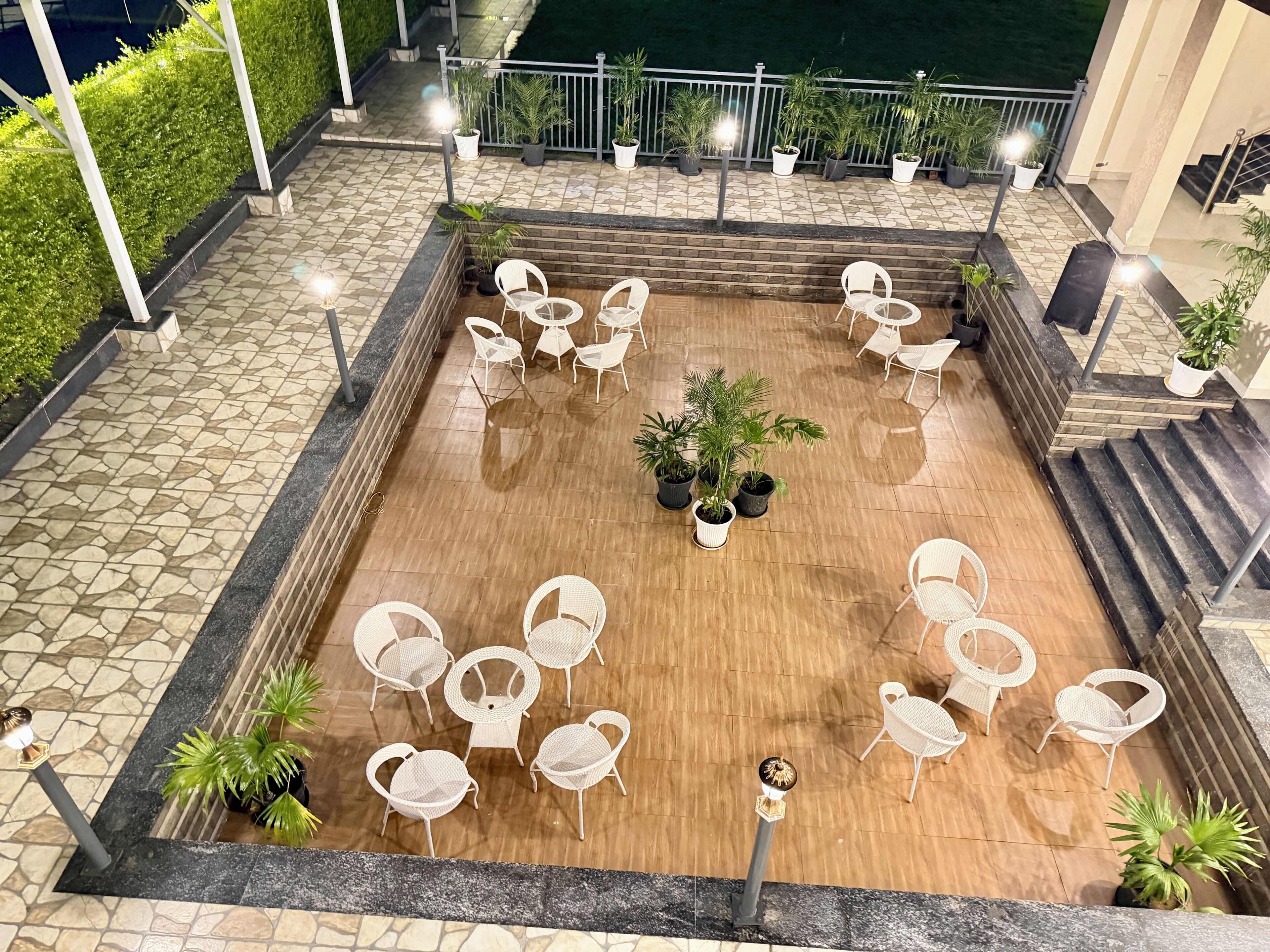 A rooftop terrace with a stone and wooden floor, surrounded by white chairs and tables, and potted plants.