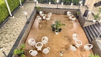 A rooftop terrace with a stone and wooden floor, surrounded by white chairs and tables, and potted plants.