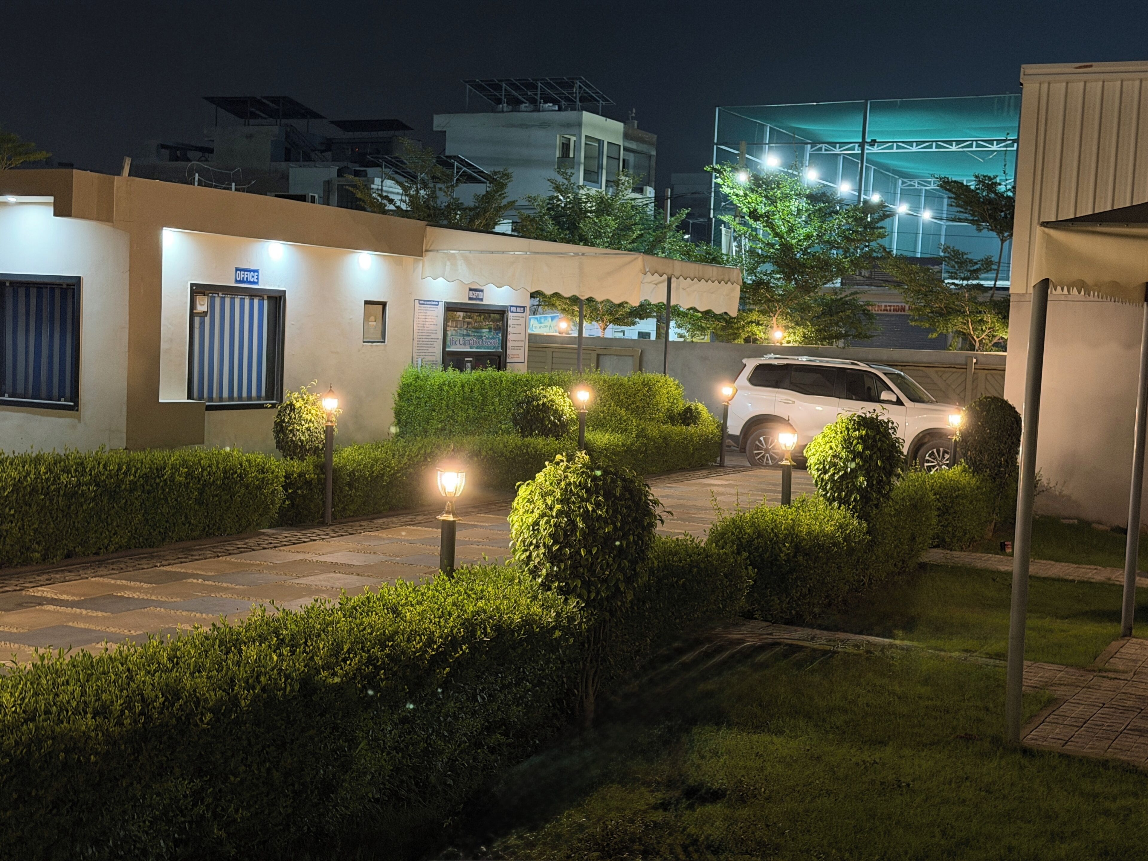 A gated residential area at night with a white SUV parked in the driveway, surrounded by well-maintained hedges and illuminated by street lamps.