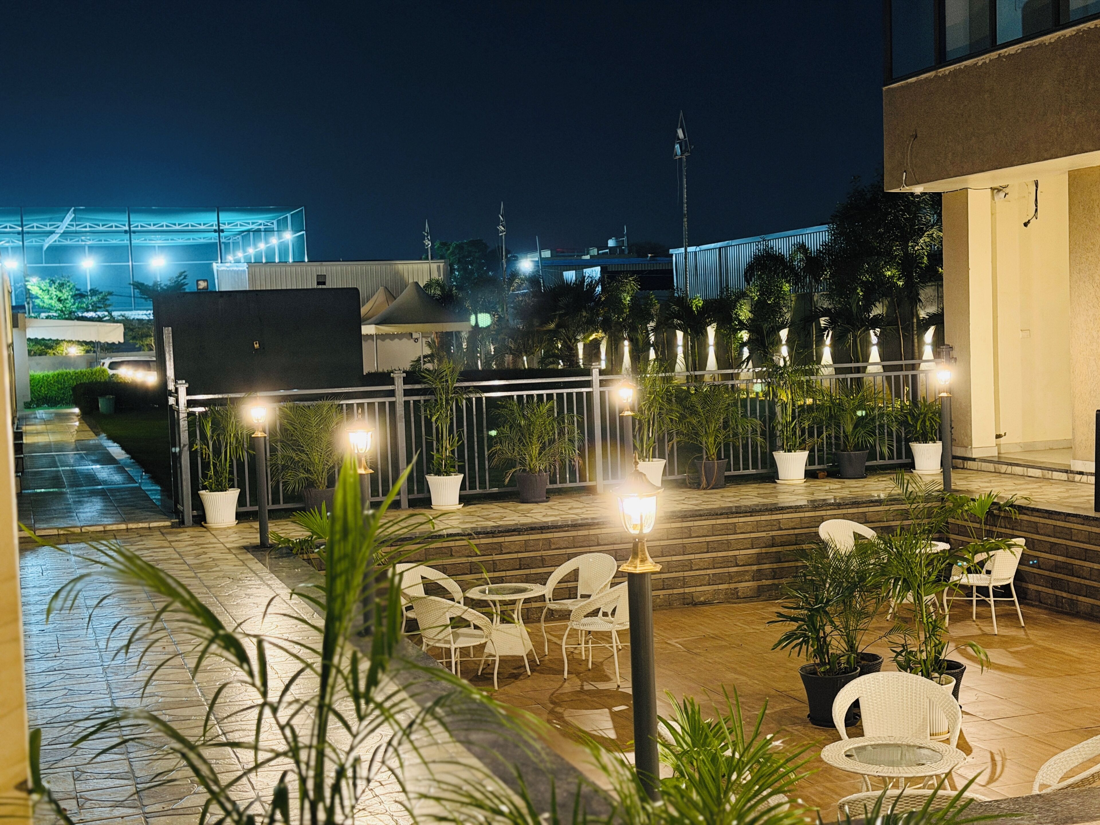 Un espace extérieur aménagé en terrasse, avec des sièges, des plantes en pot, une rambarde et une vue sur le paysage urbain illuminé la nuit.