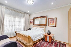 A bedroom with a bed, a bedside table, a mirror, and a window with curtains. - Aaybee Recreational Place (Midrand)