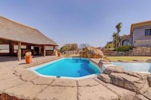 A swimming pool with a stone border, a thatched-roof structure, and a stone building with a tiled roof. - Aaybee Recreational Place (Midrand)