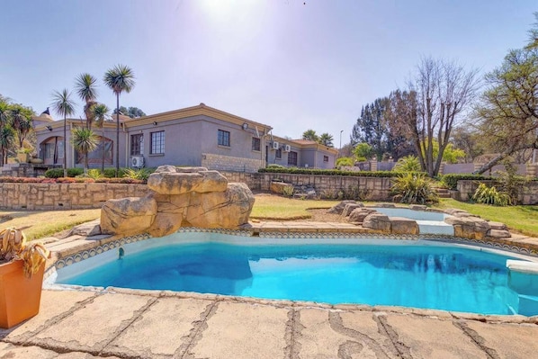 A residential property with a swimming pool, stone landscaping, and a house with a flat roof and multiple windows. - Aaybee Recreational Place (Midrand)