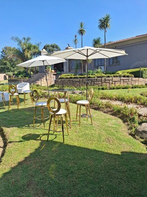 An outdoor seating area with tables and chairs, surrounded by a well-maintained garden and palm trees. - Aaybee Recreational Place (Midrand)