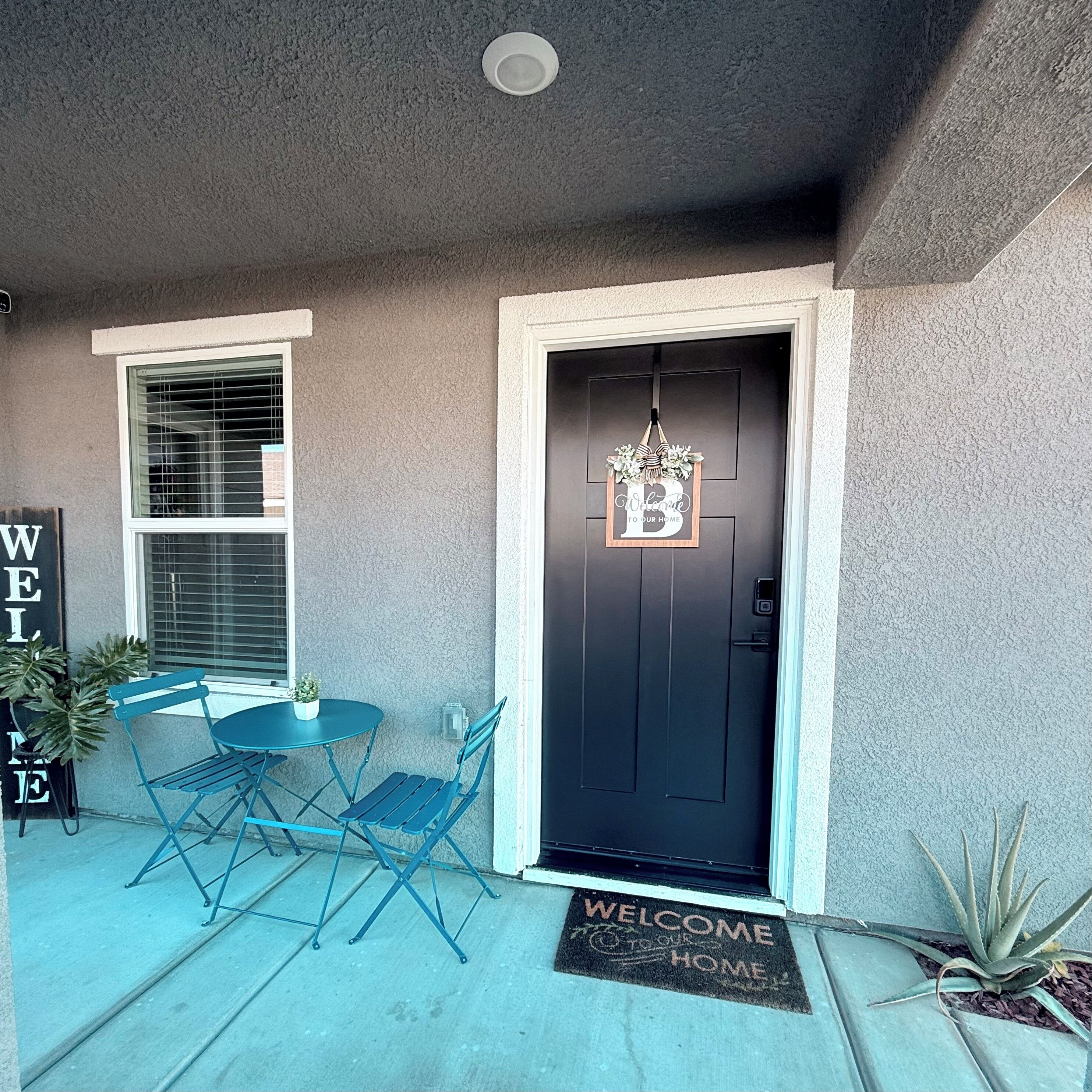 A front porch with a black door, a welcome mat, and a sign that reads 'WELCOME TO OUR HOME.'