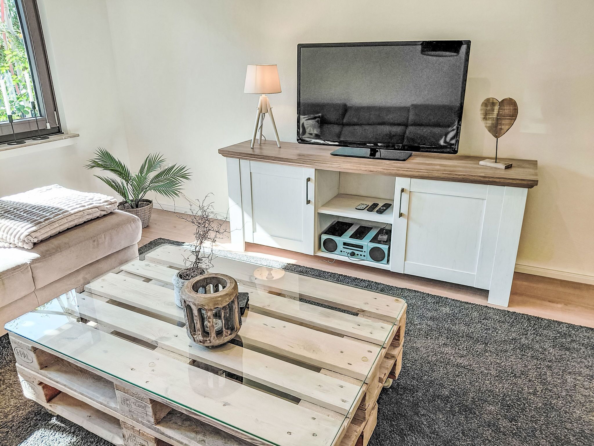 Een woonkamer met een glazen salontafel, een televisie op een houten standaard, een bank en een klein kastje met ingebouwde speakers.