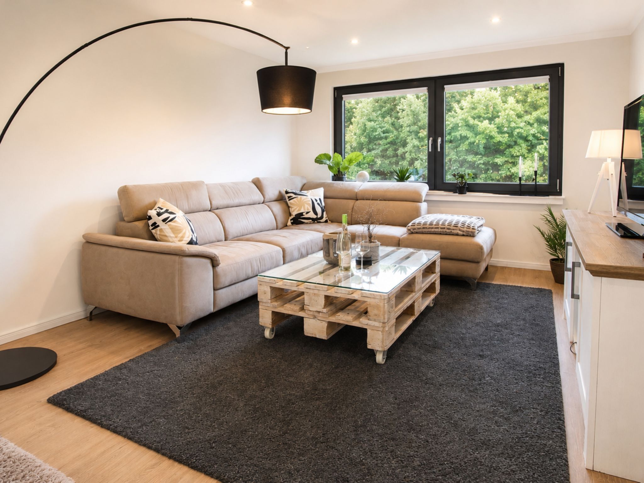 Ein modernes Wohnzimmer mit einem Ecksofa, einem Glastisch und einem großen Fenster mit Blick auf Grünflächen.