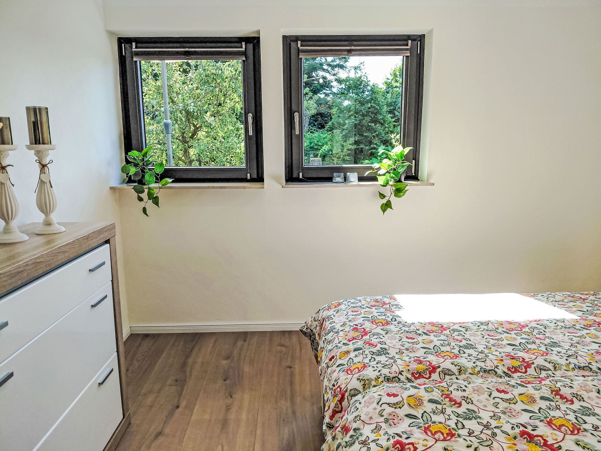Ett sovrum med två fönster, en säng med ett blommigt överkast, en byrå och en växt på fönsterbrädan.