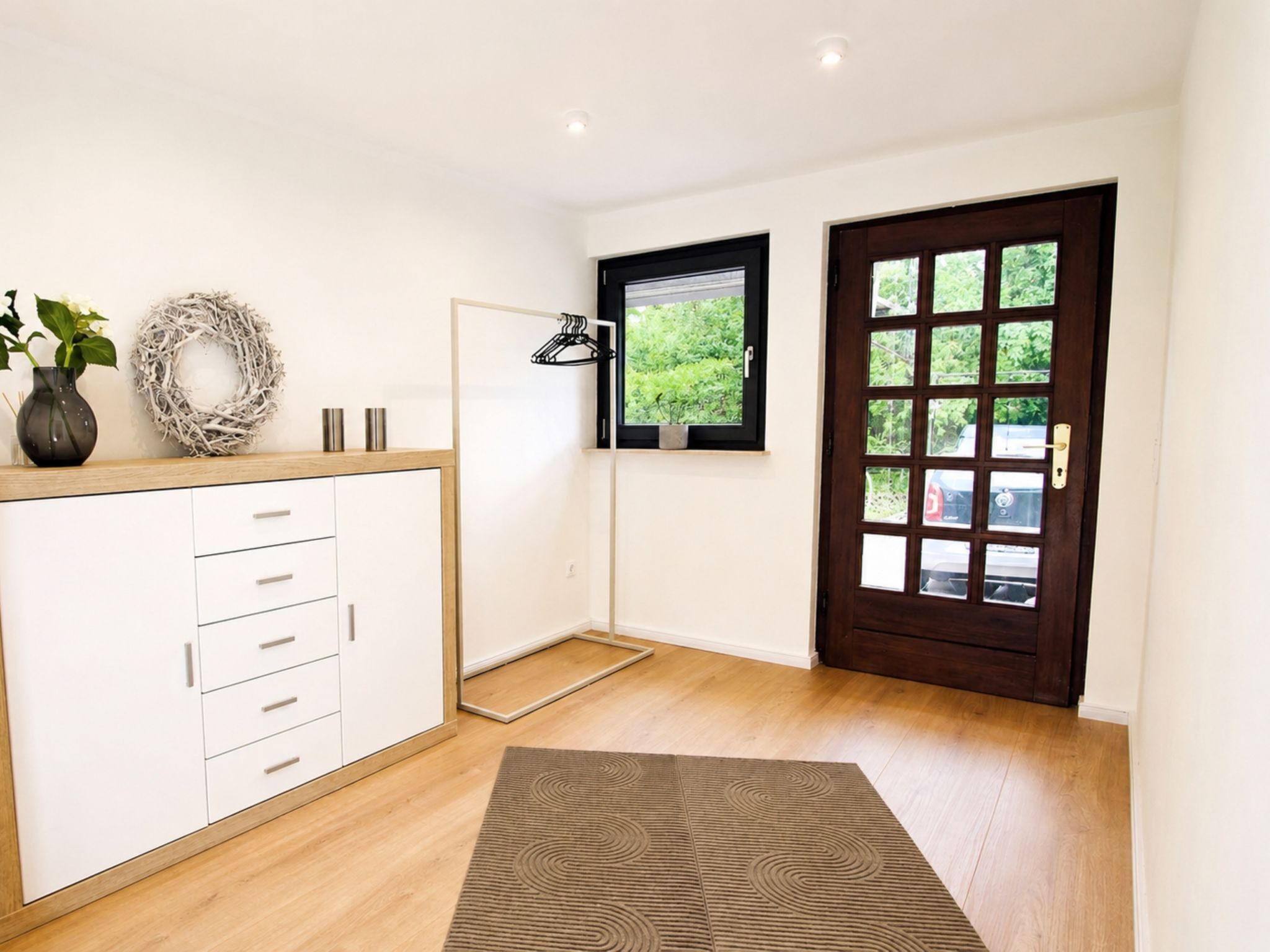 Una stanza con pavimento in legno, un armadio con cassetti, una porta con pannelli di vetro, un piccolo finestrino con vista sul verde e una ghirlanda decorativa appesa al muro.