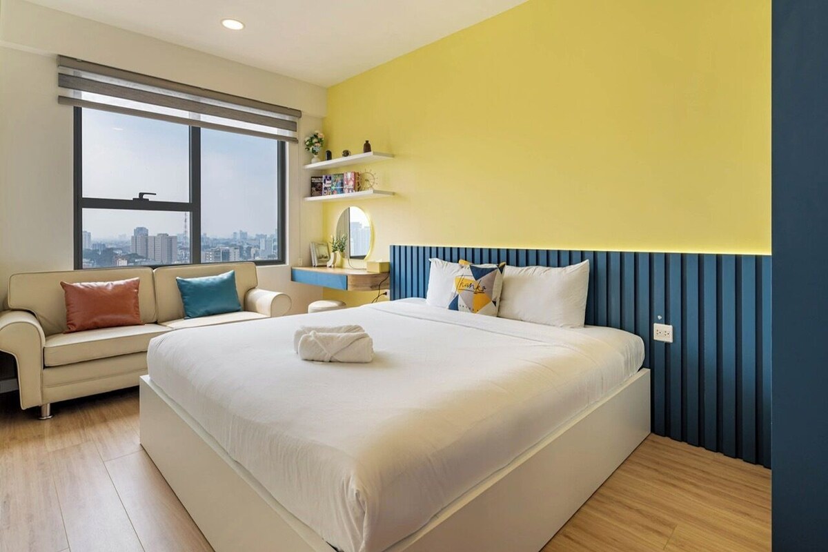 Ein Hotelzimmer mit einem großen Bett, einer Couch, Blick auf die Stadt und einem Regal mit Büchern und einem Spiegel.