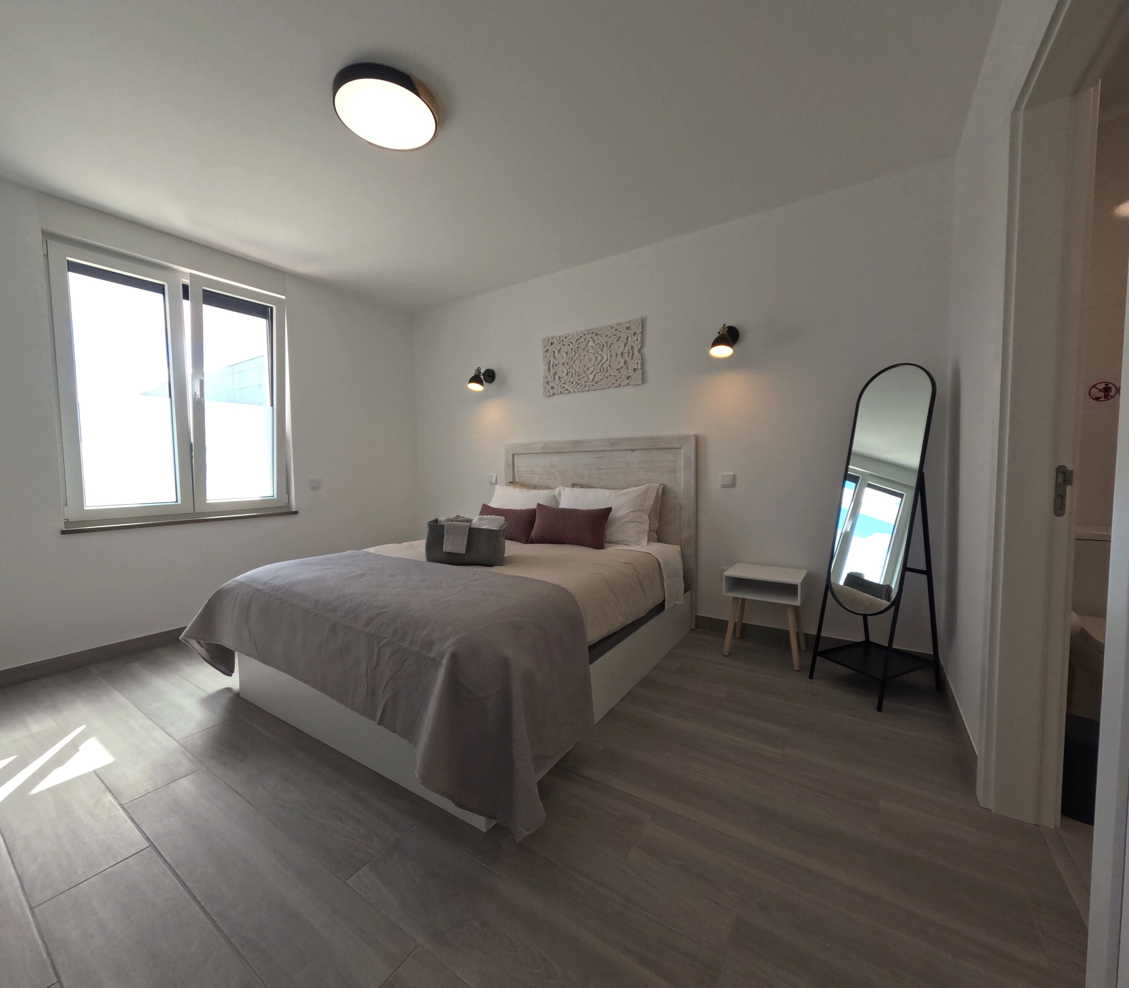 A bedroom with a bed, bedside table, mirror, and a window.