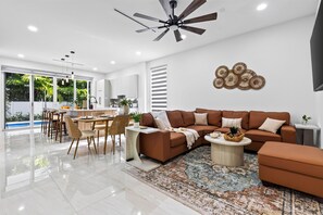 Ein modernes Wohnzimmer mit einem braunen Ecksofa, einem Couchtisch und einem Essbereich mit weißem Tisch und Stühlen.