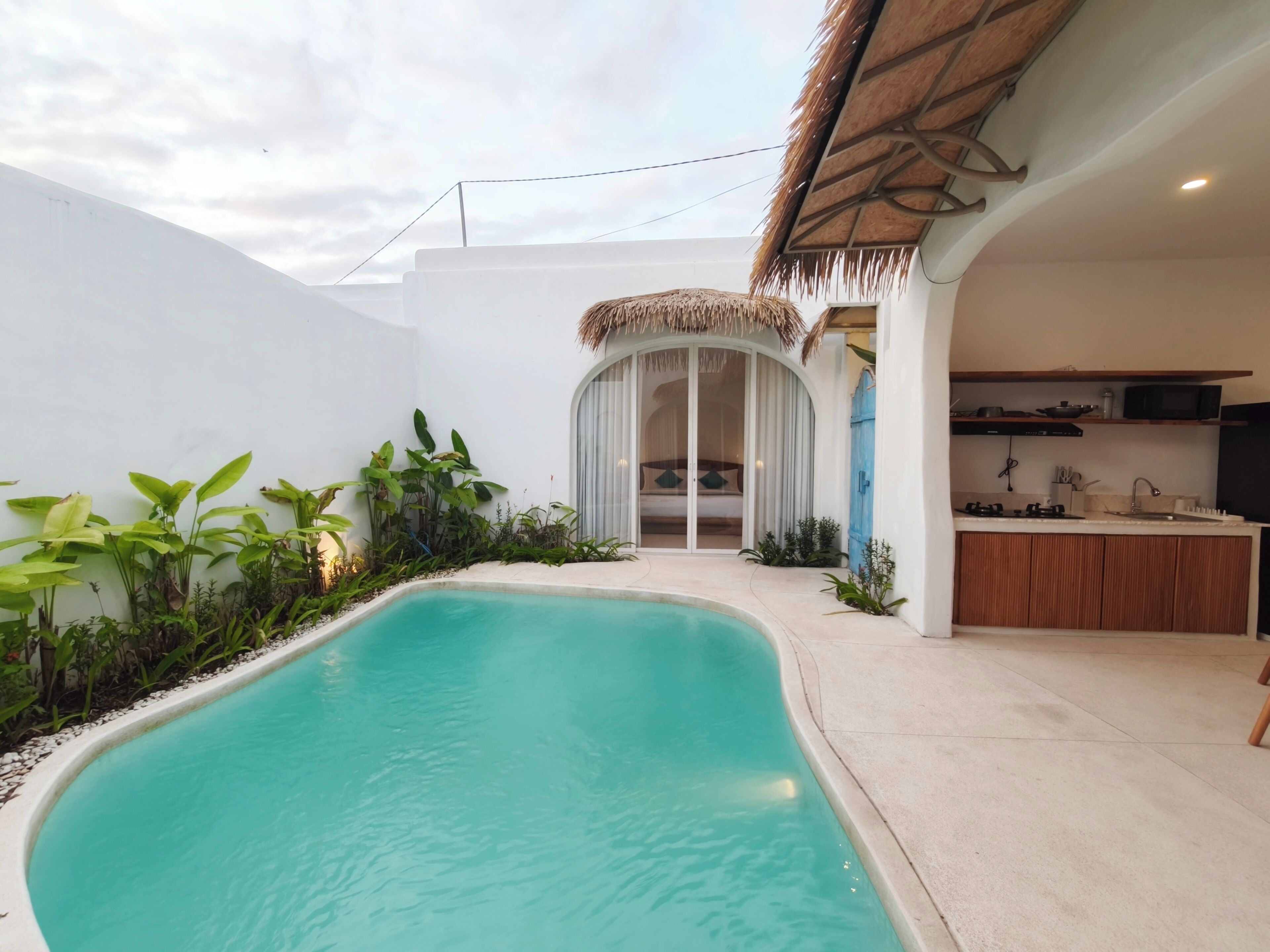 Un área de piscina con una piscina azul cristalina, rodeada de follaje y un muro blanco.