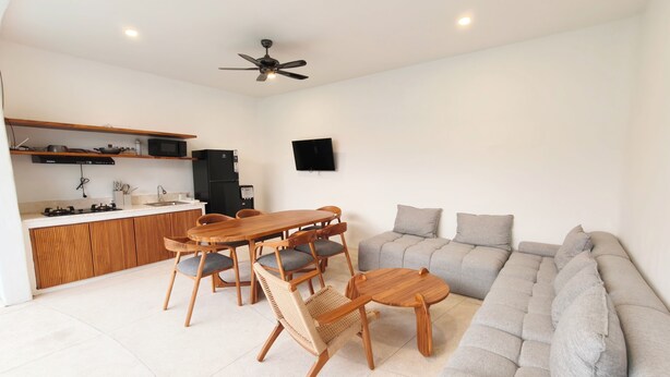 A modern living room with a sofa, a dining table with chairs, a kitchen area with a stove, and a ceiling fan. - Villa Harmony (Badung)