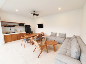 A modern living room with a sofa, a dining table with chairs, a kitchen area with a stove, and a ceiling fan. - Villa Harmony (Badung)