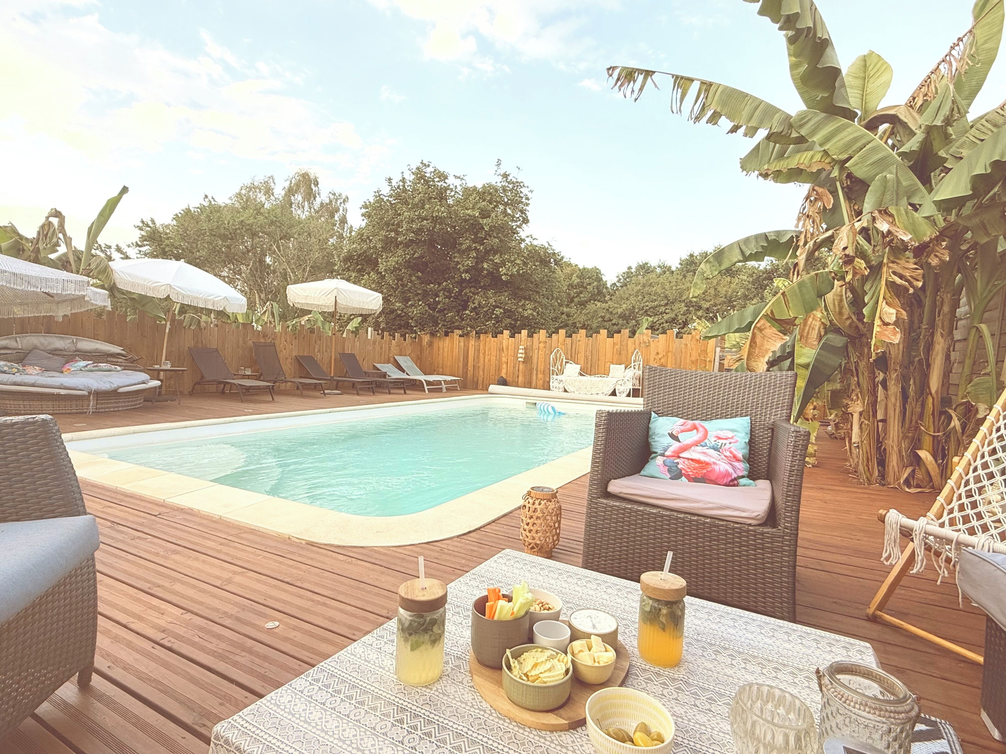 Un espace aménagé au bord de la piscine, avec des chaises longues, une table où sont servis des plats et des boissons, et une terrasse en bois.