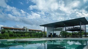 A swimming pool with a covered area, surrounded by greenery and a building in the background.