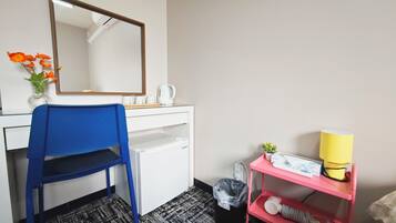 A hotel room with a desk, a blue chair, a mirror, a small plant, a yellow lamp, and a pink shelf with a hairdryer.