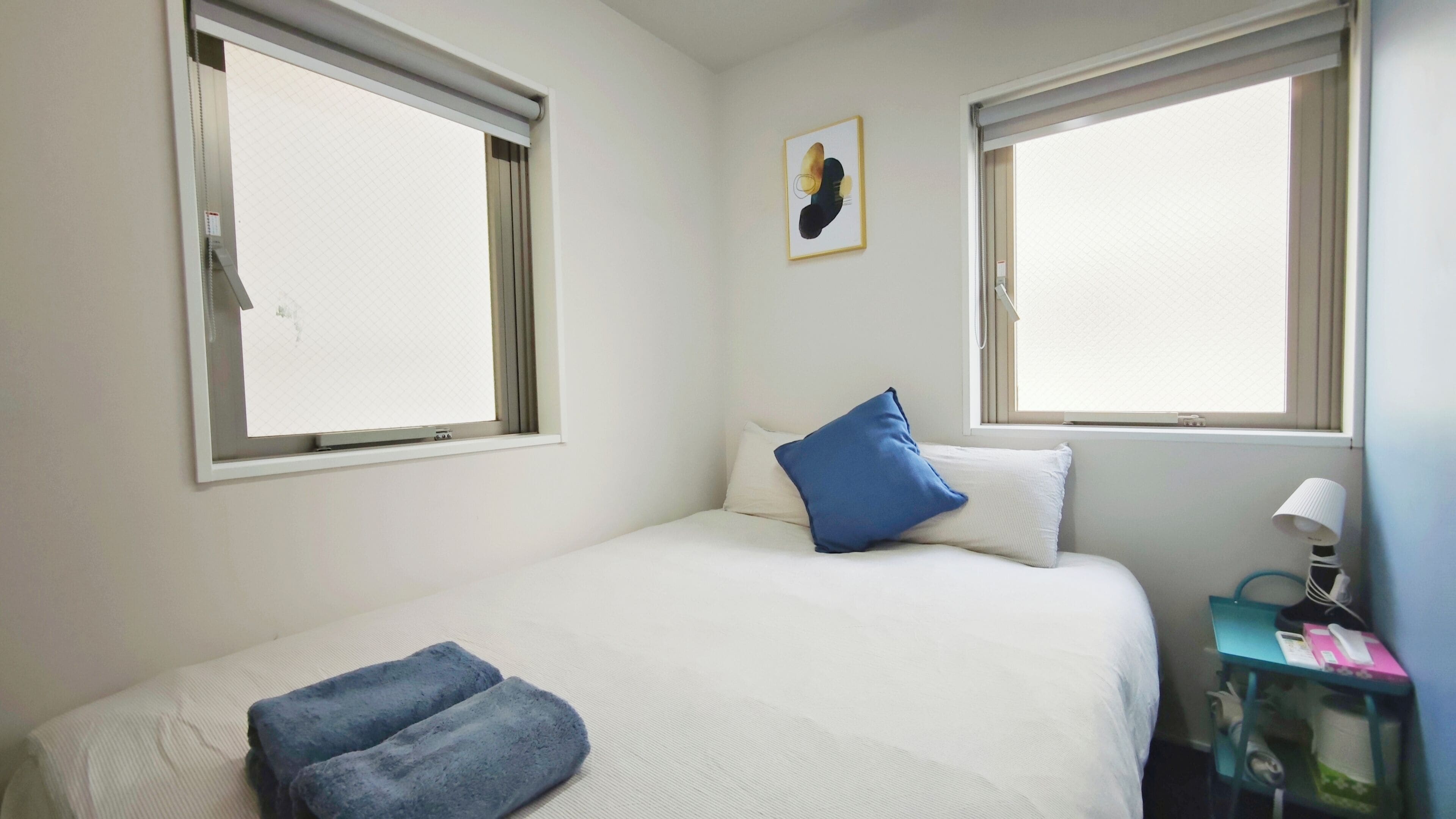 A bedroom with a bed, two windows, a painting, a bedside lamp, and a small table with a lamp.