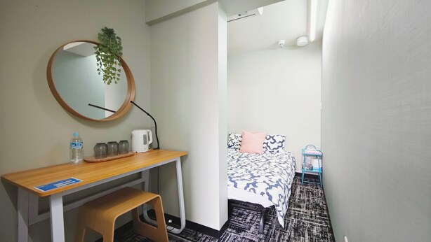 A hotel room with a wooden desk, a round mirror, a bed with patterned bedding, and a small table with a water bottle and glasses.