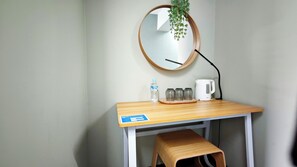 Um quarto pequeno e simples, com uma escrivaninha de madeira, um espelho, uma garrafa de água, uma chaleira branca e um banco.