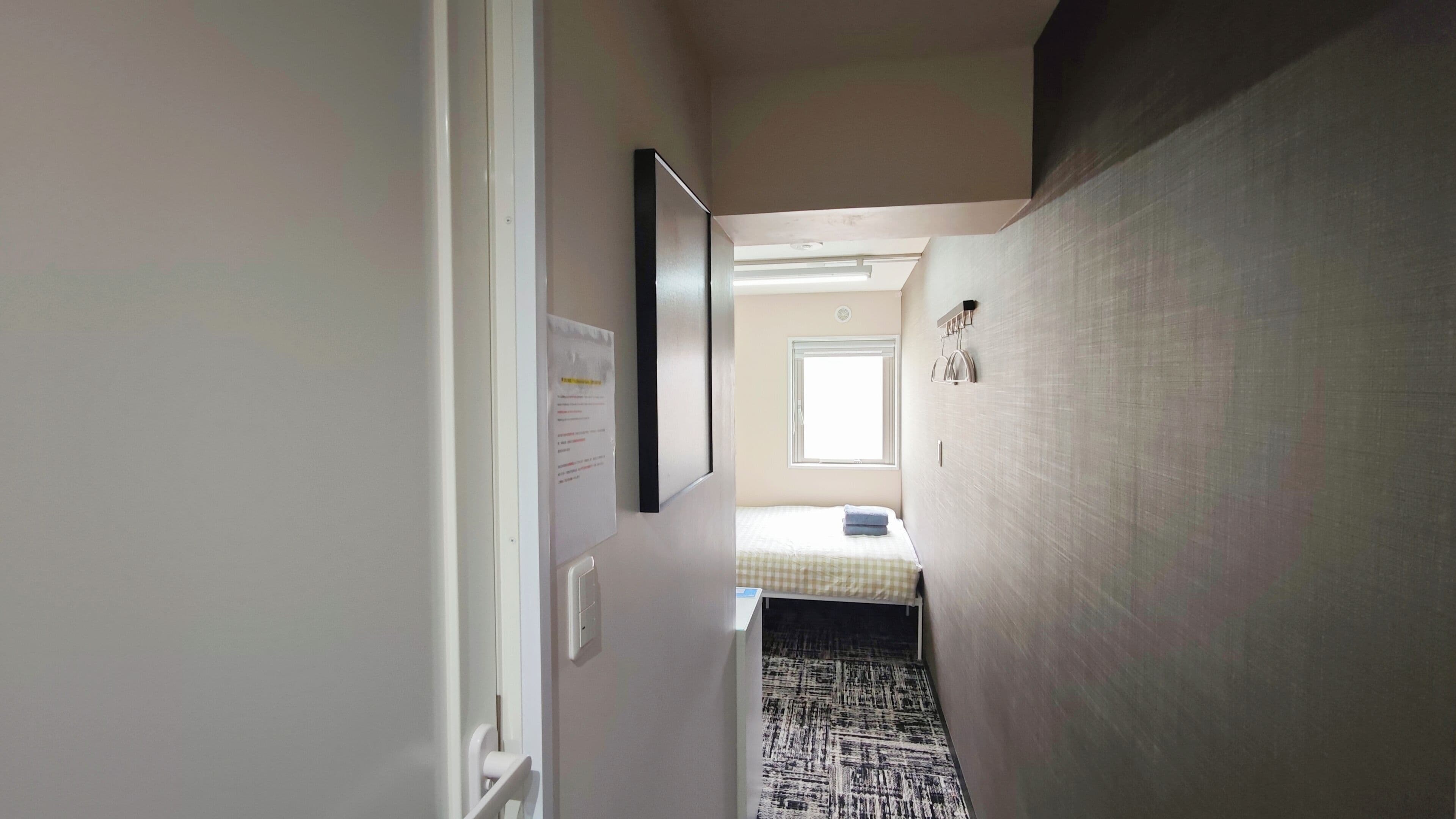 A narrow hallway with a bed at the end, a window, and a door on the left.