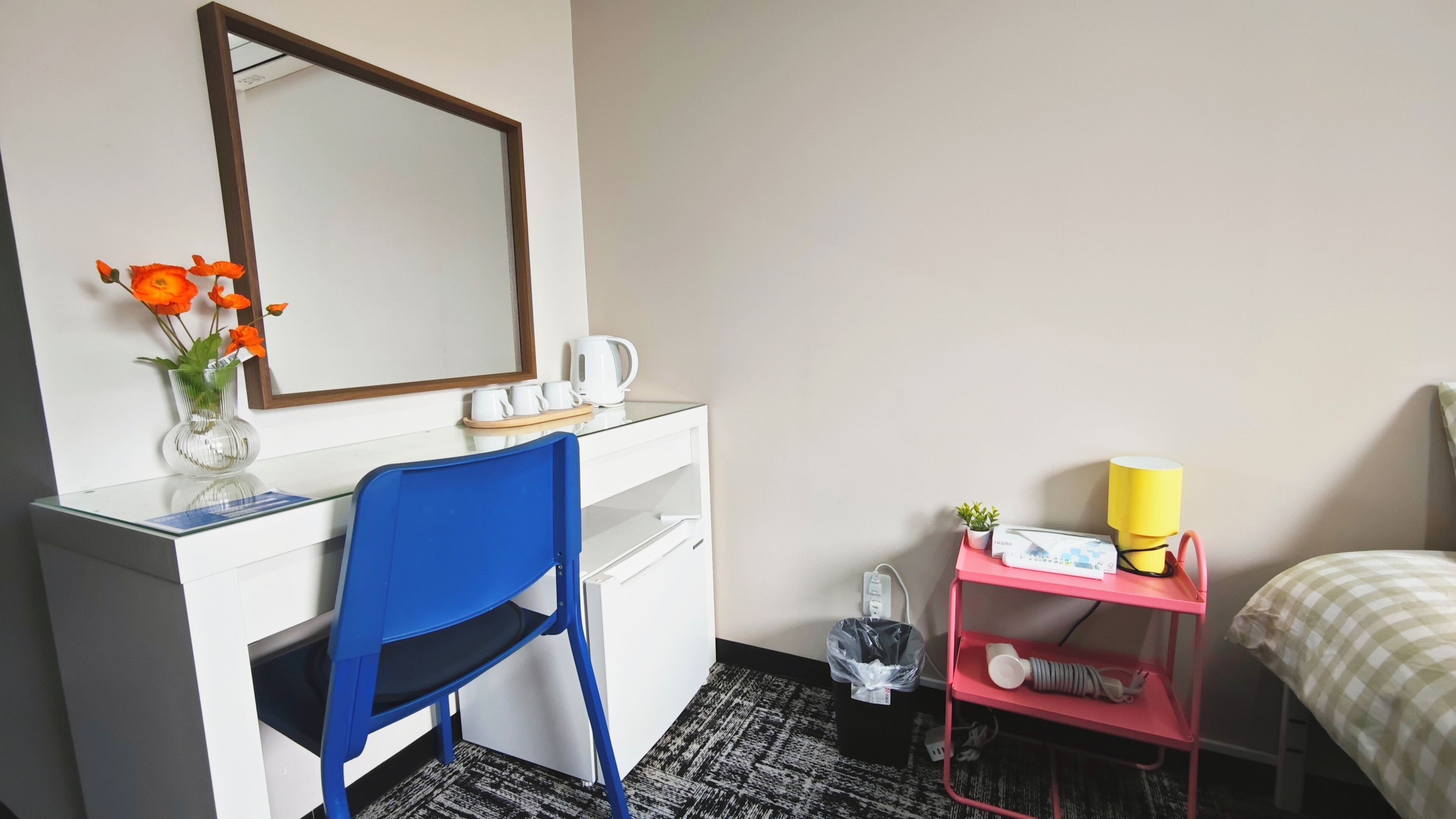 A hotel room with a desk, chair, mirror, bedside table with lamp, and a bed.