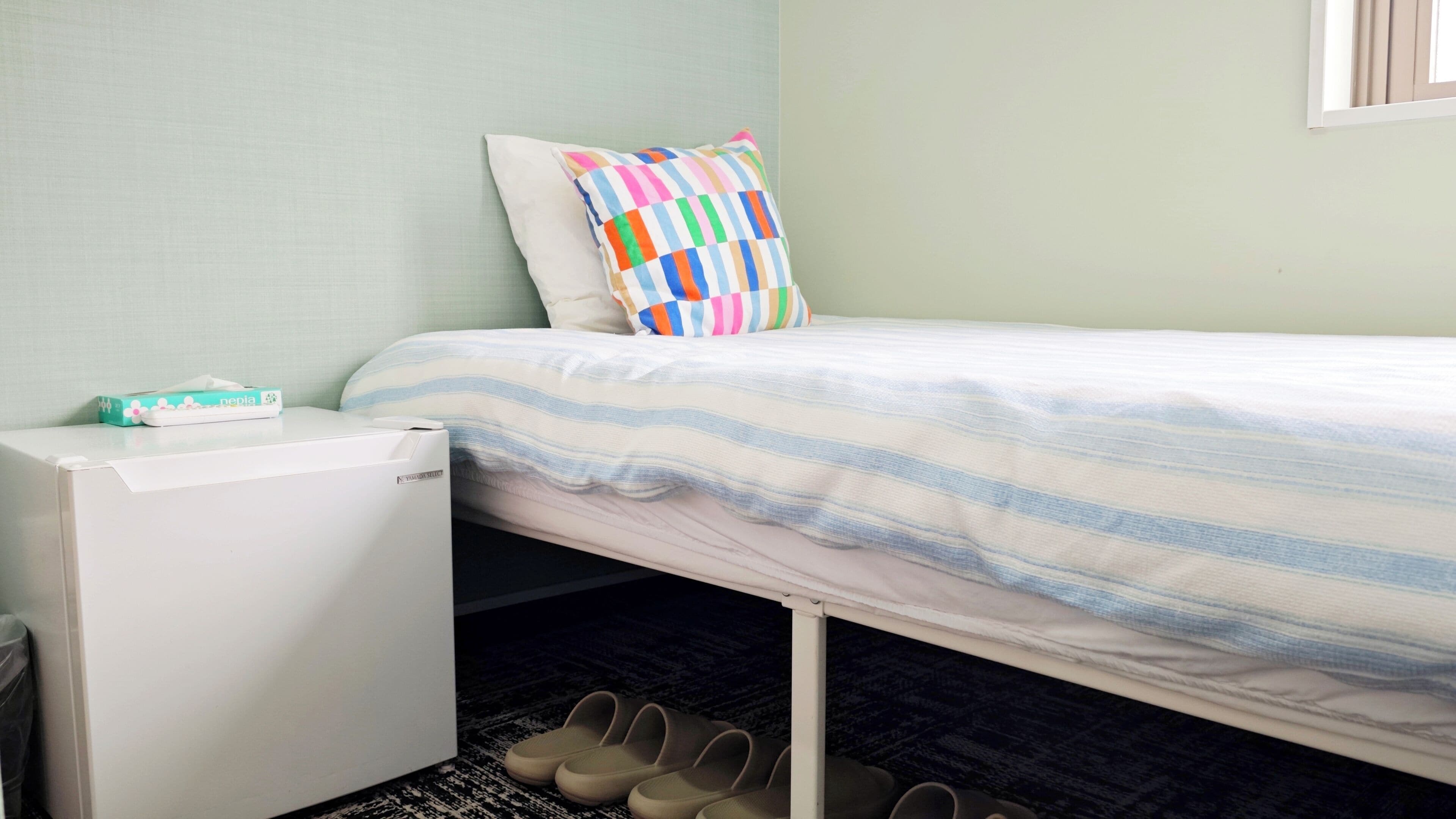 A single bed with a striped bedspread, a colorful pillow, a white bedside table with a tissue box, and a pair of shoes on the floor.