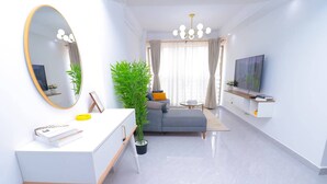 Ein modernes Wohnzimmer mit weißem Konsolentisch, einer Couch, einem großen Spiegel, einem Topf mit Pflanze, einem Fernseher und einer Kristallleuchtre.