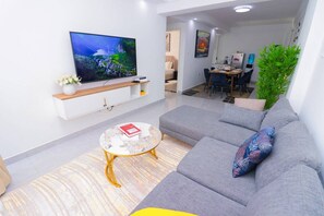 A modern living room with a flat-screen TV, a grey sofa, a coffee table, and a dining area in the background. - Goldpark Homes View Palace Apartment (Nairobi)