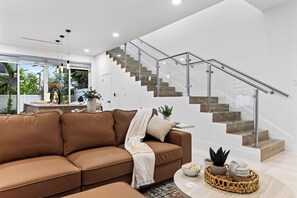 A modern living room with a brown sectional sofa, a glass staircase, and a view of the outdoors. - Shared Pool + BBQ + Private Balcony | Walk to Las Olas | Palm Perch (Fort Lauderdale)