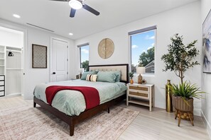 Une chambre à coucher avec un lit, des tables de chevet, un ventilateur de plafond, une fenêtre et une plante.