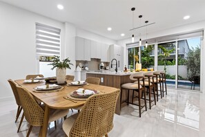 A modern kitchen with white cabinets, a central island, and a dining area with a wooden table and wicker chairs. - Shared Pool + BBQ + Private Balcony | Walk to Las Olas | Palm Perch (Fort Lauderdale)