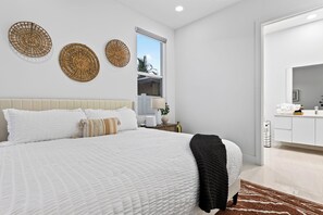A modern bedroom with a large bed, a bedside table, a window, and a bathroom visible through an open door. - Shared Pool + BBQ + Private Balcony | Walk to Las Olas | Palm Perch (Fort Lauderdale)