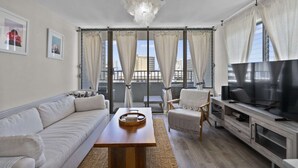 A modern living room with a sofa, a coffee table, a TV, and a balcony with curtains. - Royal Kuhio #1312 City Views (Honolulu)