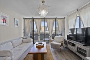 A modern living room with a sofa, a coffee table, a TV, and a balcony with curtains. - Royal Kuhio #1312 City Views (Honolulu)